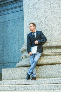 Dışarıda bekliyor. Siyah ceket, kravat, gri pantolon, deri ayakkabı, dizüstü bilgisayar, bacak bacak üstüne atma, ofis dışında ayakta duran bir iş adamı, iş sırasında mola verme.