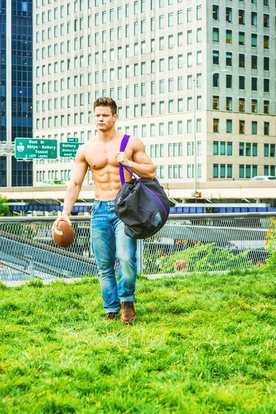 Play Hard, Work Smart. Shirtless, half naked, waring jeans, a young, strong, sexy businessman, carrying duffel bag, holding football, walking from business district in New York, having fun after work