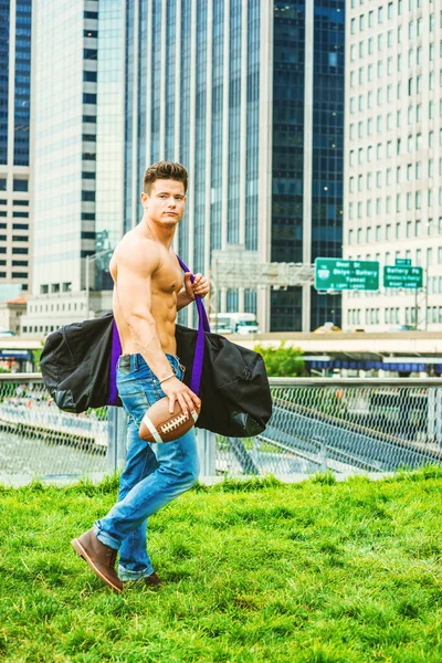 Play Hard, Work Smart. Shirtless, half naked, waring jeans, a young, strong, sexy guy, carrying big duffel bag, holding football, walking from business district in New York, going to play field.