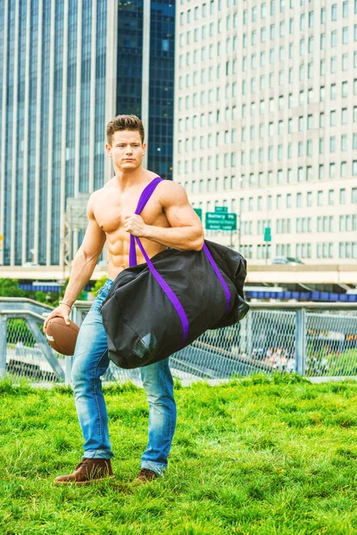Play Hard, Work Smart. Shirtless, half naked, waring jeans, a young, strong, sexy businessman, carrying duffel bag, holding football, walking from business district in New York, having fun after work