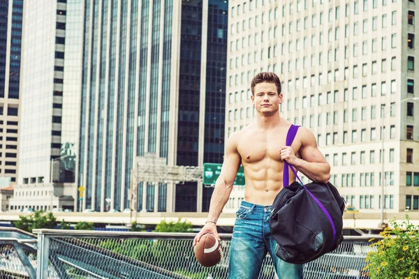 Play Hard, Work Smart. Shirtless, half naked, waring jeans, a young, strong, sexy guy, carrying big duffel bag, holding football, walking from business district in New York, going to play field.