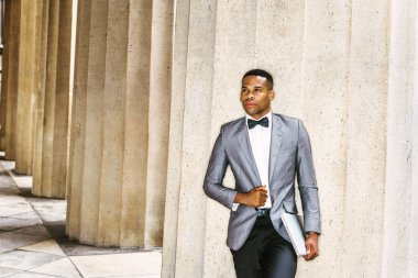 Gri ceketi, papyonu, beyaz atleti, siyah pantolonu, dizüstü bilgisayarı olan genç siyahi bir adam dışarıda durup,