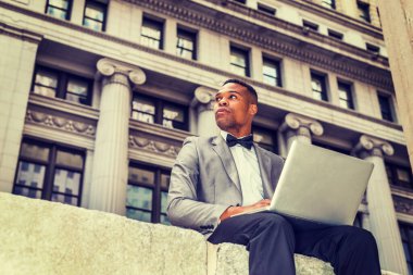 Gri ceketli, papyonlu, iş bölgesinde oturan genç, siyah bir adam, dizüstü bilgisayarla çalışıyor. Kendine güvenerek başka tarafa bakıyor. Instagram etkisi
