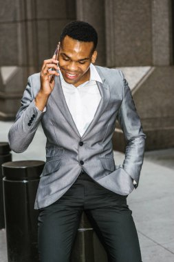 Gri ceketli, beyaz atletli, siyah pantolonlu, genç siyah bir adam sokakta oturuyor, aşağı bakıyor, gülümsüyor, telefonda konuşuyor. Şehir Hayatı.