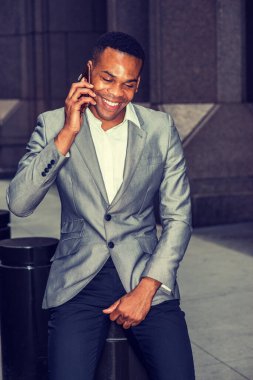 Gri ceketli, beyaz atletli, siyah pantolonlu, genç siyah bir adam sokakta oturuyor, aşağı bakıyor, gülümsüyor, cep telefonuyla konuşuyor. Instagram etkisi.