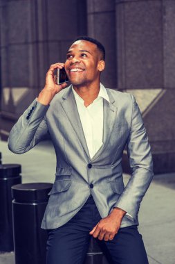Gri ceketli, beyaz atletli, siyah pantolonlu, genç siyahi bir adam sokakta oturuyor, yukarı bakıyor, gülümsüyor, telefonda konuşuyor. Şehir Günlük Yaşamı