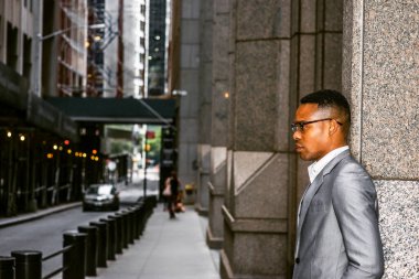 Gri ceketli, gözlüklü, sokakta dikilen siyah bir üniversite öğrencisi, üzgün, düşünceli, düşüncelere dalmış. Arka planda dar bir yolda giden bir araba