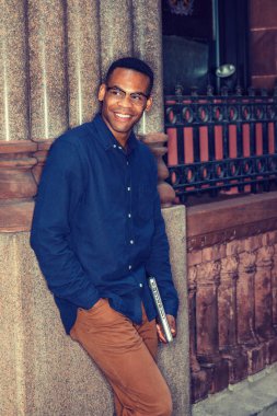 Mavi gömlekli, kahverengi pantolonlu, gözlüklü, dizüstü bilgisayarlı, genç siyah bir adam sokakta duvara yaslanmış, gülümsüyor, seni bekliyor..