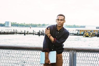 Limanda duran, mavi gömlekli, kahverengi pantolonlu, gözlüklü, bilezik takan, omuza dokunan, dizüstü bilgisayarlı siyah bir adam.