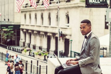 Gri ceketli, papyonlu, Wall Street 'te oturan, okuyan, dizüstü bilgisayarda çalışan, etrafa bakınan, düşünen genç siyah bir adam. Instagram etkisi.