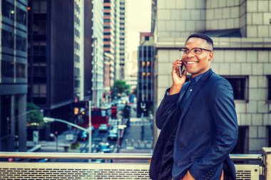 Mavi ceketli, gözlüklü, genç siyahi bir adam balkonda ayakta duruyor, caddeye bakıyor yüksek binalarla, gülümsüyor, telefonla konuşuyor..