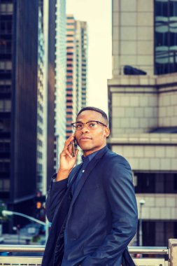 Mavi ceketli, gözlüklü, siyah bir adam balkonda ayakta duruyor, caddeye bakıyor, yüksek binalara bakıyor, telefonla konuşuyor..