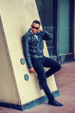 Siyah deri ceket, kot pantolon, deri ayakkabı, güneş gözlüğü, beyaz atlet, papyon, New York sokaklarında duvara yaslanan genç bir Afrikalı Amerikalı..