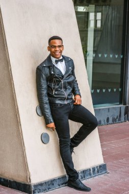 Siyah deri ceket, kot pantolon, deri ayakkabı, gözlük, beyaz atlet, papyon, New York 'ta duvarda dikilen, gülümseyen Afro-Amerikan bir adam.