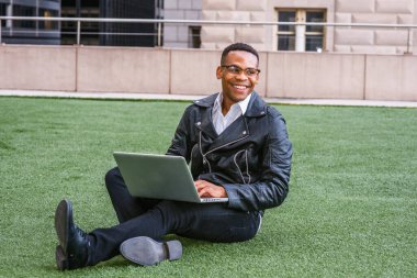 Deri ceketli, kot pantolonlu, deri ayakkabılı, gözlüklü, kampüste yeşil çimlerde oturan, dizüstü bilgisayarında çalışan, gülümseyen genç bir zenci..