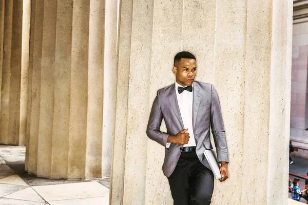 Gri ceketi, papyonu, beyaz atleti, siyah pantolonu, dizüstü bilgisayarı olan, ofisin dışındaki sütunlarda duran,