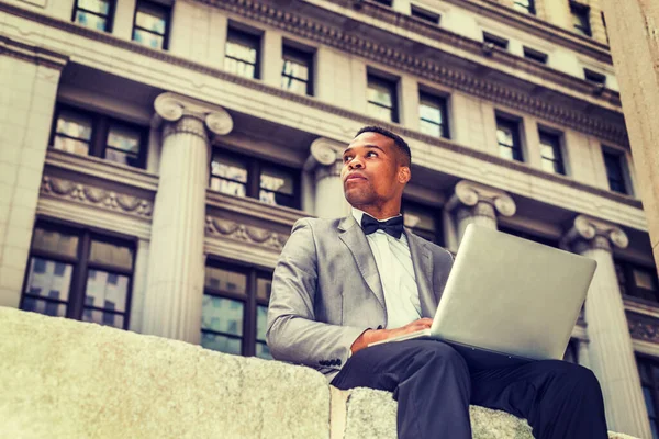 Gri ceketli, papyonlu, iş bölgesinde oturan genç, siyah bir adam, dizüstü bilgisayarla çalışıyor. Kendine güvenerek başka tarafa bakıyor. Instagram etkisi