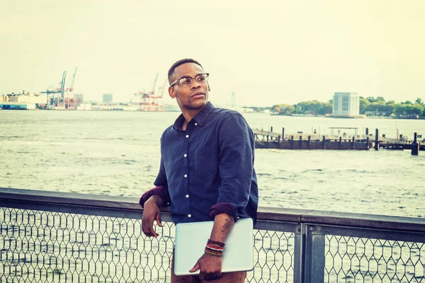 A black college student standing at harbor, wearing blue shirt, glasses, bracelets, holding laptop computer, interested, looking up.