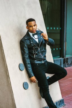 Man Urban Sonbaharı / Bahar Gelişigüzel Moda. Siyah deri ceket, beyaz atlet, siyah papyon, kot pantolon, güneş gözlüğü takan, New York sokaklarında duvara karşı duran Afrikalı Amerikalı bir adam..