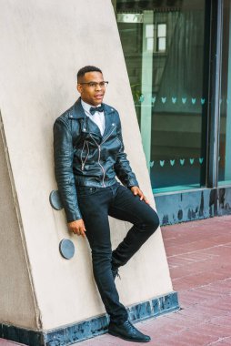 Man Urban Sonbaharı / Bahar Gelişigüzel Moda. Siyah deri ceket, beyaz atlet, siyah papyon, kot pantolon, gözlük, New York sokaklarında duvara karşı duran Afro-Amerikalı bir adam. Şehir çocuğu..