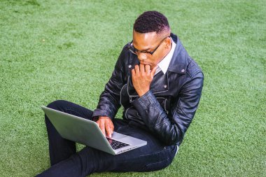 New York 'ta okuyan Afro-Amerikan öğrenci, deri ceket, kot pantolon, gözlük giyiyor, kampüste yeşil çimlerde oturuyor, dizüstü bilgisayarda çalışıyor, elleri çeneye dokunuyor, okuyor, düşünüyor..