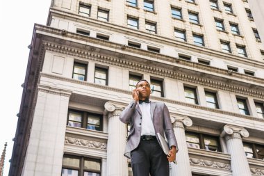 Power of Technology. African American businessman working in New York, wearing gray blazer, bow tie, standing on street, holding laptop computer, calling on cell phone. Filtered effect.