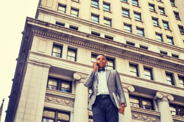 Teknolojinin gücü. New York 'ta çalışan Afro-Amerikalı işadamı, gri spor ceket, papyon takıyor, sokakta duruyor, dizüstü bilgisayarı tutuyor, cep telefonundan arıyor. Filtrelenmiş etki.