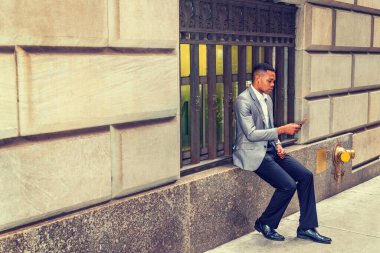 Afro-Amerikalı bir adam okuyor, sokakta cep telefonuyla mesajlaşıyor, seyahat ediyor, New York 'ta çalışıyor, gri ceket giyiyor, siyah pantolon giyiyor, deri ayakkabılar giyiyor, klasik pencere çerçevesinde oturuyor. Filtrelenmiş görünüm.