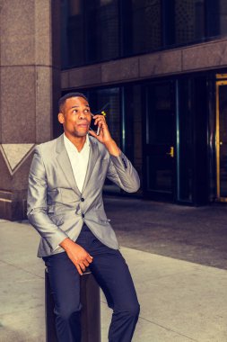 New York 'ta çalışan, gri ceketli, ofisin dışında sokakta oturan, gülümseyen, cep telefonuyla konuşan, komik suratlı, haberlerle eğlenen Afrikalı Amerikalı iş adamı. Filtrelenmiş etki.