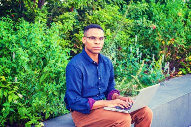 New York 'ta okuyan Afro-Amerikan üniversite öğrencisi, mavi gömlek, kahverengi pantolon, bilezik, gözlük takıyor, kampüste yeşil bitkilerin yanında oturuyor, daktilo ediyor, düşünüyor, dizüstü bilgisayarla çalışıyor..