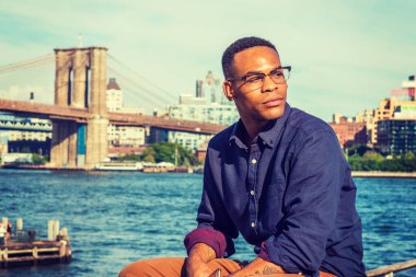 Afrikalı Amerikalı seyahat ediyor, New York 'ta çalışıyor. Mavi gömlekli, kahverengi pantolonlu, gözlüklü, East River 'da oturan, etrafa bakınan, seni bekleyen bir üniversite öğrencisi. Brooklyn Köprüsü arka planda.