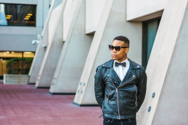 Man Urban Sonbaharı / Bahar Gelişigüzel Moda. Siyah deri ceket, siyah kot pantolon, güneş gözlüğü, beyaz atlet, siyah papyon, New York 'ta sokakta duran genç bir Afrikalı Amerikalı. Şehir çocuğu..