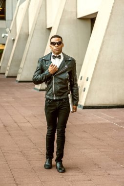 Man Urban Sonbaharı / Bahar Gelişigüzel Moda. Siyah deri ceket, beyaz atlet, siyah papyon, kot pantolon, güneş gözlüğü, dizüstü bilgisayar taşıyan, New York 'ta sokakta yürüyen Afrikalı Amerikalı bir adam..