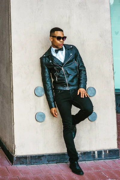 Man Urban Sonbaharı / Bahar Gelişigüzel Moda. Siyah deri ceket, beyaz atlet, siyah papyon, kot pantolon, güneş gözlüğü, New York sokaklarında duvara karşı duran Afrikalı Amerikalı adam..