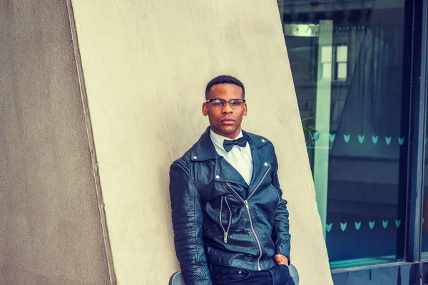 Man Urban Sonbaharı / Bahar Gelişigüzel Moda. Siyah deri ceket, beyaz atlet, siyah papyon, güneş gözlüğü, New York sokaklarında duvara karşı duran Afro-Amerikalı bir adam. Filtrelenmiş etki.