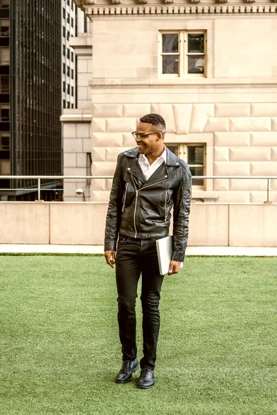 New York 'ta okuyan Afro-Amerikan üniversite öğrencisi, siyah deri ceket, kot pantolon, deri ayakkabı, gözlük, dizüstü bilgisayar, kampüste yeşil çimlerde yürümek, gülümsemek..