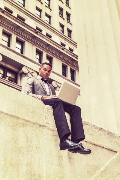 New York 'ta çalışan, gri ceket, papyon, siyah pantolon, deri ayakkabı giyen, sokakta oturan, okuyan, dizüstü bilgisayarda çalışan, düşünen Afrikalı Amerikalı iş adamı. Filtrelenmiş etki.