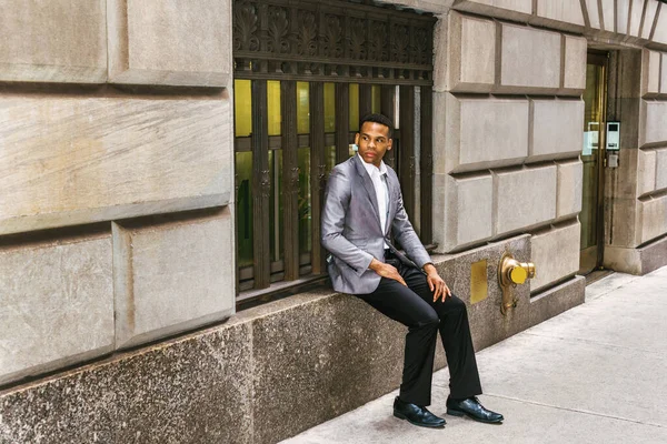 Afro-Amerikalı bir adam seyahat ediyor, New York 'ta çalışıyor, gri ceket giyiyor, siyah pantolon giyiyor, deri ayakkabılar giyiyor, klasik pencere çerçevesinde oturuyor, başka tarafa bakıyor, düşünüyor. Filtrelenmiş görünüm.