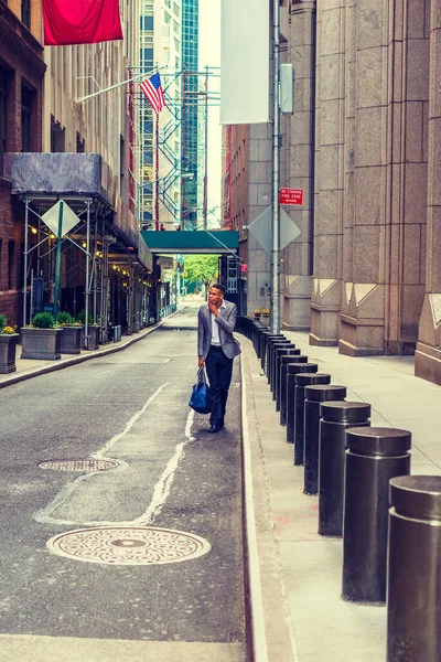 African American Businessman traveling, working in New York. Young black man walking on narrow old street with high buildings, carrying blue bag, wearing blazer, hand touching chin, looking around