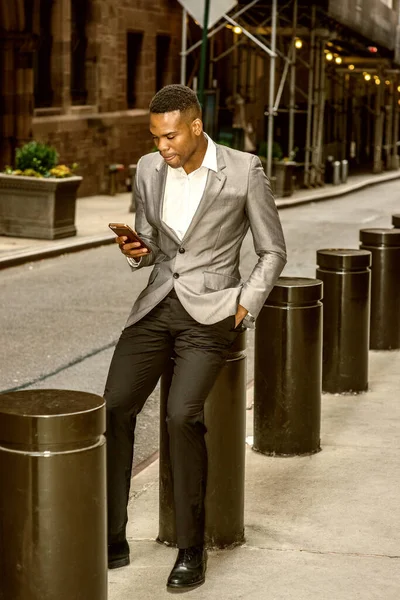 New York 'ta sokakta mesajlaşan, gri ceket, beyaz atlet, siyah pantolon, deri ayakkabı giyen, metal sütuna oturan, cep telefonu tutan, dil ısıran, mesajlar okuyan Afro-Amerikan bir adam..