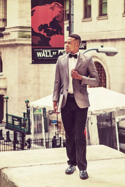 New York 'ta çalışan, gri ceketli, papyonlu, siyah pantolonlu, ayakkabılı, dizüstü bilgisayarlı, Wall Street' te yumruğunu göğsüne dayamış bir Afro-Amerikalı iş adamı. Retro filtrelenmiş etkisi.