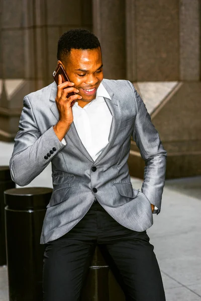 Happy African American Businessman working in New York. Wearing gray blazer, white undershirt, black pants, young black man sitting on street outside office, smiling, talking on phone. City Daily Life.