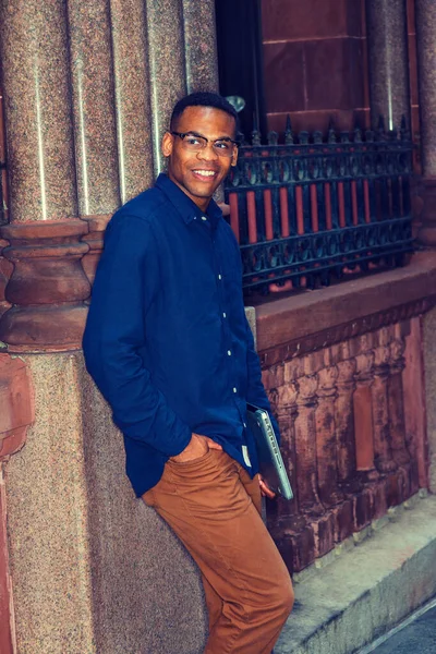 New York 'ta okuyan Afro-Amerikan üniversite öğrencisi, mavi gömlekli, kahverengi pantolonlu, gözlüklü, dizüstü bilgisayarlı, klasik tarzda duvarın karşısında duran, gülümseyen, seni bekleyen.