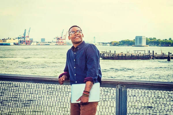 African American businessman traveling on East River in New York.  A black college student standing at harbor, wearing blue shirt, glasses, bracelets, holding laptop computer, interested, looking up.