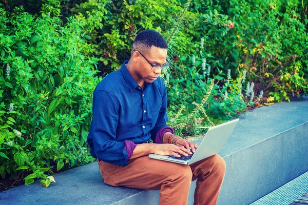 New York 'ta okuyan Afro-Amerikan üniversite öğrencisi, mavi gömlek, kahverengi pantolon, bilezik, gözlük takıyor, kampüste yeşil bitkilerin yanında oturuyor, aşağı bakıyor, daktilo ediyor, dizüstü bilgisayarla çalışıyor..