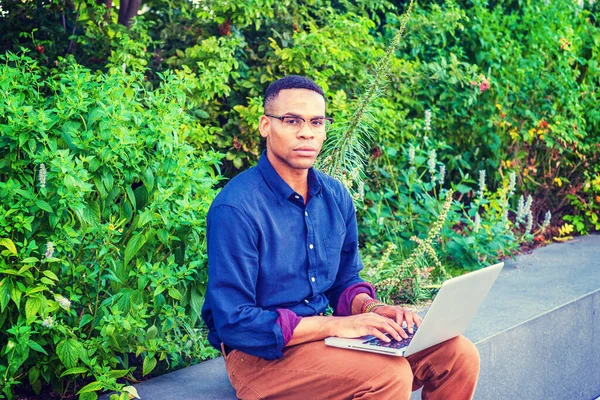 New York 'ta okuyan Afro-Amerikan üniversite öğrencisi, mavi gömlek, kahverengi pantolon, bilezik, gözlük takıyor, kampüste yeşil bitkilerin yanında oturuyor, daktilo ediyor, düşünüyor, dizüstü bilgisayarla çalışıyor..