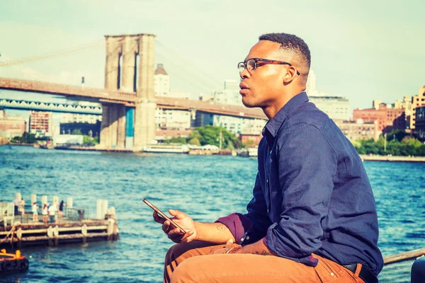 African American Man traveling in New York. Wearing blue shirt, brown pants, glasses, bracelets, a college student sitting at harbor, checking message on mobile phone, thinking. Bridge on background.