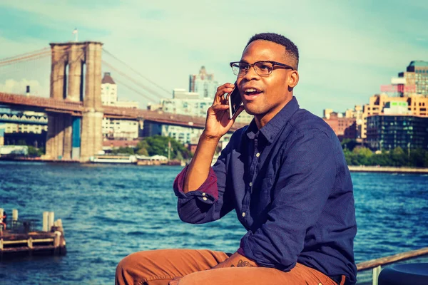 Afrikalı Amerikalı seyahat ediyor, New York 'ta çalışıyor. Mavi gömlekli, kahverengi pantolonlu, gözlüklü, East River 'da oturan, gülümseyen, telefonla konuşan bir üniversite öğrencisi. Brooklyn Köprüsü arka planda.