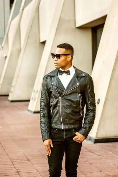 Man Urban Sonbaharı / Bahar Gelişigüzel Moda. Siyah deri ceket, siyah kot pantolon, güneş gözlüğü, beyaz atlet, siyah papyon, New York 'ta sokakta duran genç bir Afrikalı Amerikalı. Şehir çocuğu..