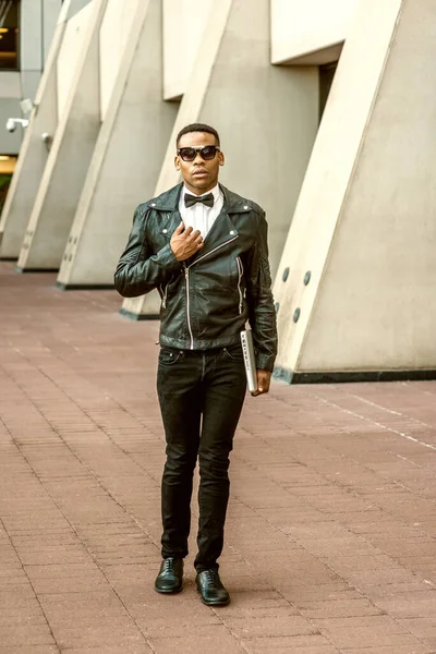 Man Urban Sonbaharı / Bahar Gelişigüzel Moda. Siyah deri ceket, beyaz atlet, siyah papyon, kot pantolon, güneş gözlüğü, dizüstü bilgisayar taşıyan, New York 'ta sokakta yürüyen Afrikalı Amerikalı bir adam..